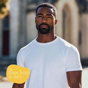 May include: A man wearing a white crew neck t-shirt. The image includes the text "a mockup a day" in a yellow oval. The background is a blurred outdoor setting with a building.