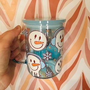 Könnte beinhalten: Eine klare Glas-Tasse mit einem blauen Deckel und einem Schneemann-Design. Die Tasse hat einen Henkel und ist mit mehreren Schneemännern verziert, die Ohrenschützer und Schneeflocken tragen.