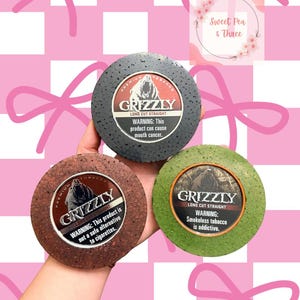Puede incluir: Tres latas de tabaco Grizzly Long Cut Straight en gris, marrón y verde. Cada lata tiene una etiqueta de advertencia y el logotipo de Grizzly con un oso. El fondo es un patrón de cuadros rosas y blancos con lazos rosas.