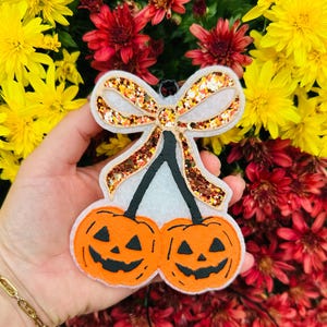 May include: A Halloween-themed ornament featuring two orange jack-o'-lanterns with black smiling faces, connected by black stems to a white bow with gold glitter accents. The ornament is held against a backdrop of yellow and red flowers.
