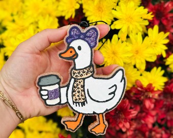May include: A white goose-shaped ornament with a purple bow, leopard print scarf, and a coffee cup. The goose has orange feet and beak. The background is a mix of yellow and red flowers.