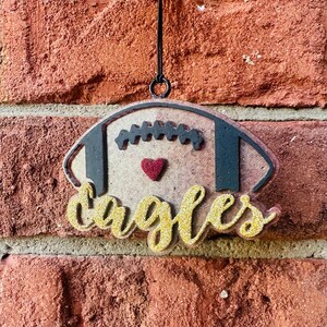 Peut inclure: Une décoration de football en bois avec le mot "eagles" en lettres pailletées dorées. Le ballon de football est peint dans un motif brun et noir avec un cœur rouge au centre.