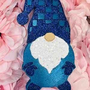 May include: A blue gnome ornament with a glittery finish. The gnome has a white beard, a tan nose, and a blue hat with a checkered pattern. The ornament is hanging against a backdrop of pink flowers.
