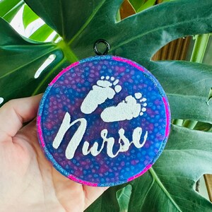 May include: A round, blue and pink resin ornament with white glitter. The ornament has a white outline and a white inscription that reads "Nurse". There are two white footprints in the center of the ornament.