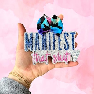 May include: A silver glitter keychain with the text "MANIFEST that shit" in blue and pink glitter. A blue and purple bow is attached to the top of the keychain.