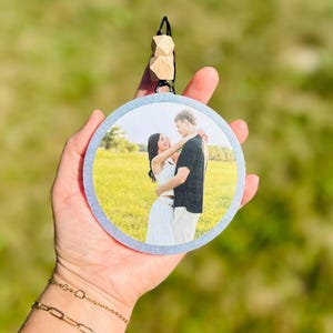 May include: A round, personalized photo ornament featuring a couple embracing in a field. The ornament has a light blue glittery border and is suspended by a black string with wooden beads. The background is a blurred green.