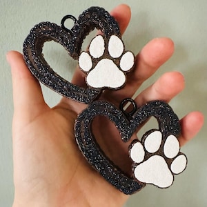 May include: Two black glitter heart-shaped ornaments with white paw prints. The ornaments have a loop at the top for hanging.