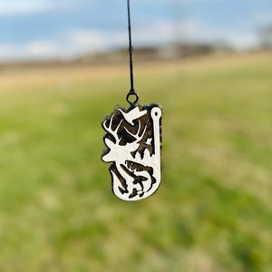 May include: A white silhouette of a deer, fish, and a fishing hook on a black and brown camouflage background. The ornament is hanging from a black string.