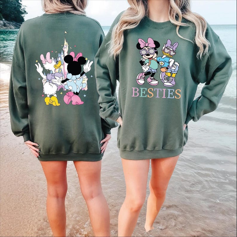 Two women wearing matching green sweatshirts with Minnie Mouse and Besties printed on them, standing on a beach.