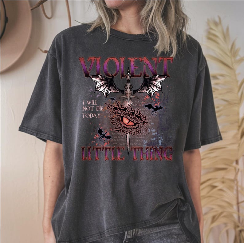 A person wearing a black t-shirt with a graphic design featuring a dragon, bats, and text that reads "I will not die today" and "Little thing".
