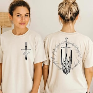 May include: A cream-colored short-sleeve t-shirt featuring a black graphic print. The front displays a small sword with a ring around it. The back features a larger graphic of a sword, a detailed helmet, and circular text in a script font.