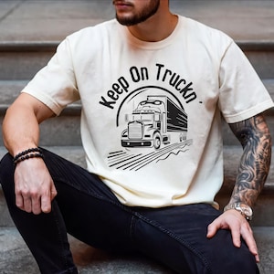May include: A cream-colored short-sleeve t-shirt featuring a black graphic of a semi-truck and the text "Keep On Truckn" arched above the vehicle. The design includes stylized motion lines beneath the truck, creating a sense of forward movement.
