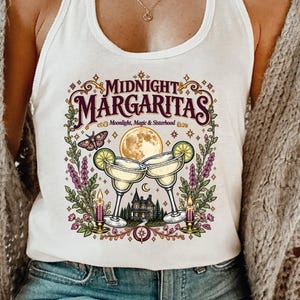May include: A white tank top featuring a graphic print with the text "MIDNIGHT MARGARITAS" and "Moonlight, Magic & Sisterhood". The design includes two margaritas, a full moon, a moth, two lit candles, and a house surrounded by lavender sprigs.