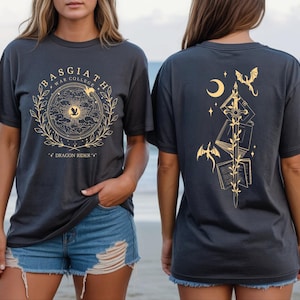 two women wearing black t-shirts with gold designs on them. The t-shirts have a dragon and moon motif, and the women are standing on a beach with the ocean in the background.