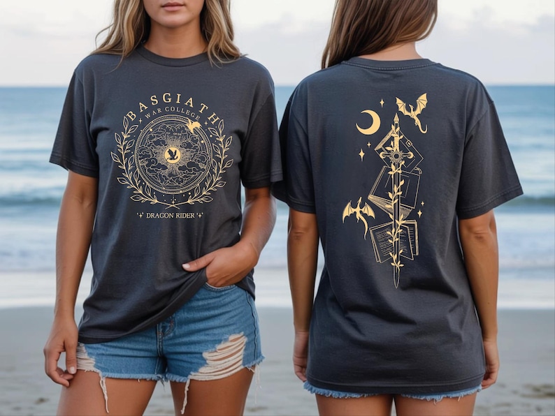 two women wearing black t-shirts with gold designs on them. The t-shirts have a dragon and moon motif, and the women are standing on a beach with the ocean in the background.