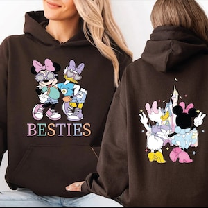 A woman wearing a brown hoodie with a graphic of Minnie Mouse and Donald Duck on the front, and the text "BESTIES" on the back.