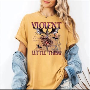 A woman wearing a yellow t-shirt with a graphic design featuring a dragon, a red eye, and the text "VIOLENT LITTLE THING".