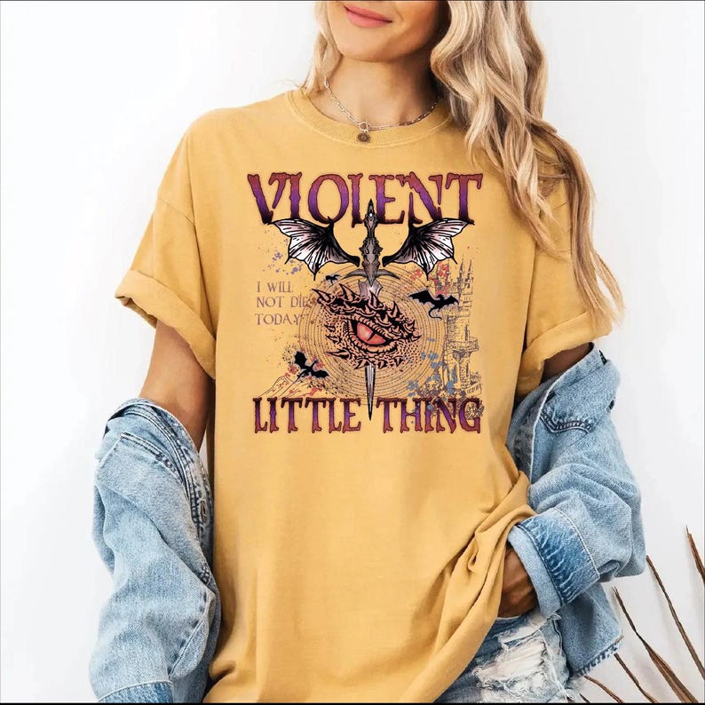 A woman wearing a yellow t-shirt with a graphic design featuring a dragon, a red eye, and the text "VIOLENT LITTLE THING".