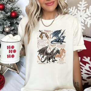 A woman wearing a white t-shirt with a graphic design of four dragons, including a dragon with a blue body and a dragon with a red body. She is holding a mug with the phrase "HO HO" written on it.