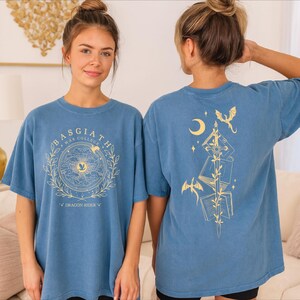 A woman wearing a blue t-shirt with a dragon design and text on it, standing in a room with a couch and a gold wall decoration.