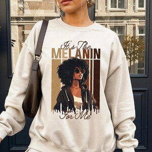 May include: Cream-colored sweatshirt with the text "It's The MELANIN For Me" and a graphic of a Black woman wearing sunglasses. The graphic features shades of brown and black. The sweatshirt has long sleeves and a crew neck.