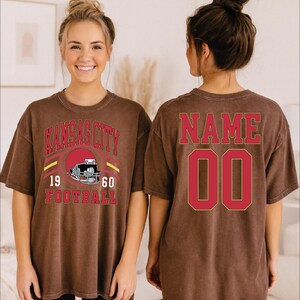 A woman wearing a brown t-shirt with a Kansas City football design and the text "Name 00" printed on the back.