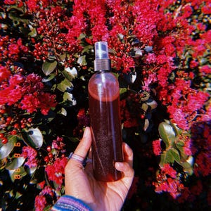 May include: A clear glass bottle with a spray nozzle filled with a dark red liquid. The bottle is being held in front of a background of pink flowers.
