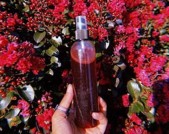May include: A clear glass bottle with a spray nozzle filled with a dark red liquid. The bottle is being held in front of a background of pink flowers.