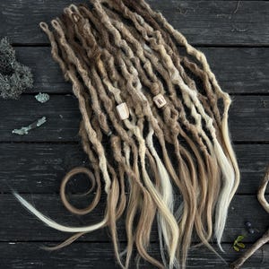 Puede incluir: Un conjunto de rastas hechas a mano en tonos marrones, rubios y blancos. Las rastas están dispuestas en forma de abanico, con algunos mechones rizados. Se adjuntan dos pequeñas cuentas de madera. Se muestra sobre una superficie de madera oscura.