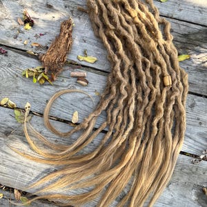 May include: A bundle of light brown dreadlocks with a wavy texture, lying on a weathered wooden surface. The dreadlocks have a natural, sun-kissed appearance, with lighter tips. A wooden bead is attached to the dreadlocks. Scattered leaves and twigs are around the dreadlocks.