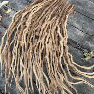 Peut inclure: Ensemble de dreadlocks faits à la main, dans des tons bruns et blonds. Les dreads sont disposés sur une surface en bois, avec les extrémités des cheveux passant à un blond plus clair. La texture semble naturelle et légèrement ondulée.