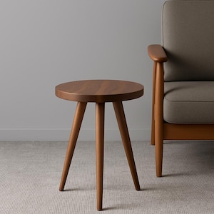 May include: A small, round wooden side table with three angled legs. The table has a dark brown finish and is placed on a light-coloured carpet. A grey armchair is partially visible in the background.