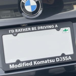 May include: Black car with a license plate frame that reads "I'D RATHER BE DRIVING A" and "Modified Komatsu D355A". The BMW logo is visible above the license plate.