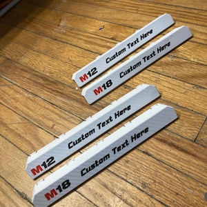 May include: Four white, elongated, rectangular plastic battery tool mounts with beveled edges. Two are labelled M12 and two are labelled M18 in red and black text. Each mount features the black printed text Custom Text Here on the top surface.