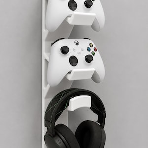 May include: A white vertical wall mount organizer featuring three integrated hooks. The top two hooks hold white wireless game controllers with black joysticks and colored buttons. The bottom hook holds a black gaming headset with green stitching.