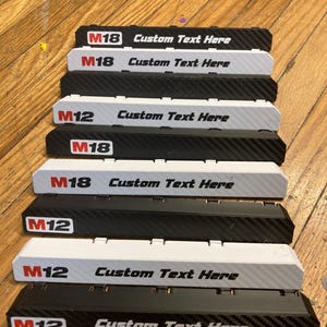 Puede incluir: Una colección de soportes rectangulares para baterías en blanco y negro, con texturas de fibra de carbono y acabados sólidos. Cada soporte muestra la marca M12 o M18 y el texto Custom Text Here, diseñado para organizar baterías.