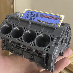 May include: A grey, 3D-printed engine block with five cylindrical openings. A business card with the text "print THAT! Winnipeg based" is inserted into the top. The engine block is held in a hand.