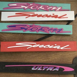 以下が含まれることがあります： 「Storm」「Special」「Ultra」の文字がレトロフォントでデザインされた長方形のキーホルダーのコレクション。 ターコイズ、白、赤、黒など様々な色があり、ピンクと紫のアクセントが施されています。 各キーホルダーにはコードが付いています。