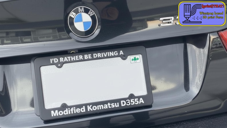 Puede incluir: Coche negro con un marco de matr&iacute;cula que dice "I'D RATHER BE DRIVING A" y "Modified Komatsu D355A". El logotipo de BMW es visible encima de la matr&iacute;cula.