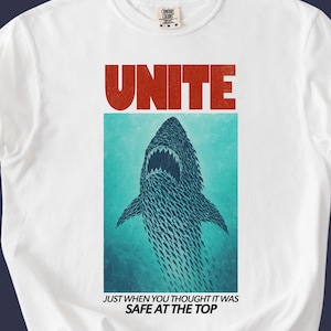 以下が含まれることがあります： 白いTシャツ。赤い文字で「UNITE」と書かれ、サメのグラフィックが描かれています。サメの下には「JUST WHEN YOU THOUGHT IT WAS SAFE AT THE TOP」というテキストがあります。クルーネックと半袖です。