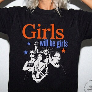 May include: Black t-shirt with the text "Girls will be girls" in orange and blue. The shirt features a graphic of several women, including the Statue of Liberty and Ruth Bader Ginsburg. The shirt is from Rebel & Roots Co.