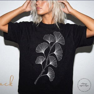 Pode incluir: T-shirt preta com um design de folhas de ginkgo brancas estilizadas. A camisa tem mangas curtas e gola redonda. A palavra "Black" está escrita em itálico na parte inferior esquerda. A marca "Rebel & Roots Co." está no canto superior direito.