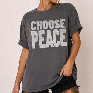 May include: A charcoal gray short-sleeve t-shirt with a distressed white graphic print on the front that reads CHOOSE PEACE in bold, capital letters. The garment features a crew neckline and a relaxed, loose-fitting silhouette with rolled sleeves.