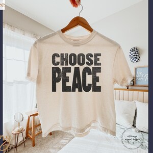 May include: An ivory-colored short-sleeve t-shirt featuring the text CHOOSE PEACE in a bold, distressed black font centered on the chest. The garment has a crew neckline and a boxy, relaxed fit, hanging from a wooden hanger against a bright background.