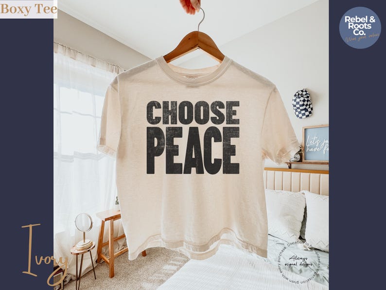 May include: An ivory-colored short-sleeve t-shirt featuring the text CHOOSE PEACE in a bold, distressed black font centered on the chest. The garment has a crew neckline and a boxy, relaxed fit, hanging from a wooden hanger against a bright background.