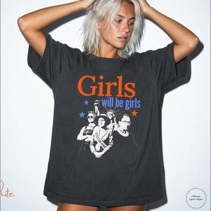 May include: Dark gray t-shirt with the text "Girls will be girls" in orange and blue, featuring a graphic of women. The shirt is a relaxed fit and has short sleeves. The image includes the "Rebel & Roots Co." logo.