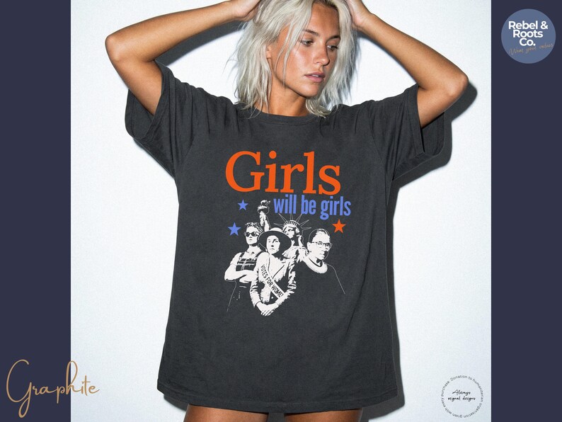 May include: Dark gray t-shirt with the text "Girls will be girls" in orange and blue, featuring a graphic of women. The shirt is a relaxed fit and has short sleeves. The image includes the "Rebel & Roots Co." logo.