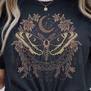 May include: Black t-shirt featuring a detailed moth design with a crescent moon, stars, and floral elements. The moth has intricate wing patterns in gold and brown, surrounded by a circular design with botanical accents. A gold necklace is visible.