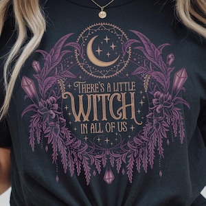 May include: Dark gray t-shirt with a graphic design. The design features a crescent moon, stars, and the text "There's a Little Witch in All of Us" in a decorative font, surrounded by purple floral and crystal elements.