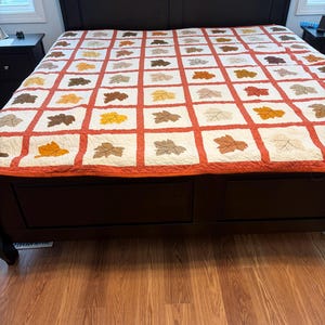 May include: A dark brown bed with a leaf-patterned quilt. The quilt features squares with leaf designs in shades of brown, orange, and yellow, set against a white background with red borders. The bed frame has a headboard and two drawers.